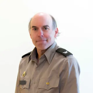 Person in a gray uniform shirt with shoulder insignia faces the camera against a plain white background.