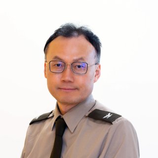Person wearing glasses, a gray uniform shirt, and a black tie poses against a plain light background.