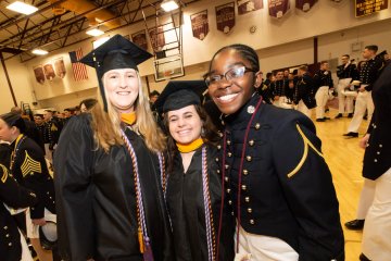 Three people wearing graduation caps and gowns standing next to a person in a dark uniform with gold trim in a gym.