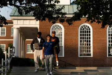 Three students walk down campus steps in front of a brick Chapel building with tall arched windows.
