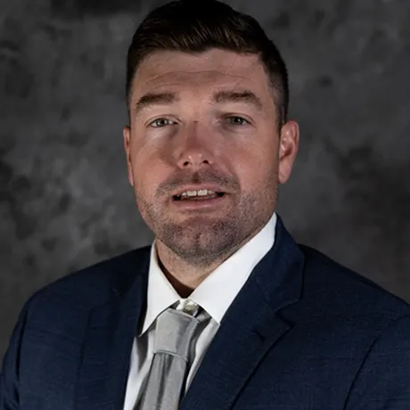 A man with short dark brown hair, wearing a navy blue suit, white shirt and gray tie, standing against a textured dark gray background.