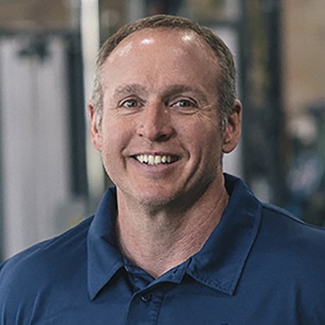 A smiling man with short light brown hair wearing a blue polo shirt stands in a fitness facility with workout equipment in the blurred background.