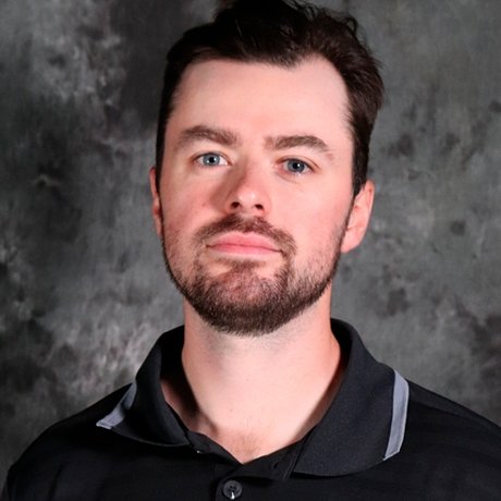 A man with short dark hair and a trimmed beard, wearing a black collared shirt with gray accents, standing against a dark, marbled background.