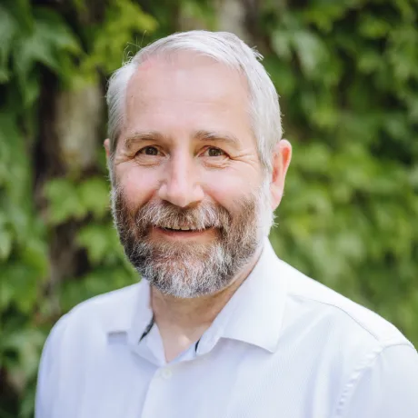 Man in a light blue shirt, smiling in front of foliage.
