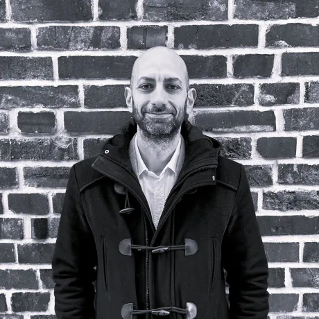 Man in a dark coat standing in front of a brick wall.