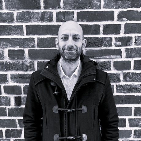 Man in a dark coat standing in front of a brick wall.