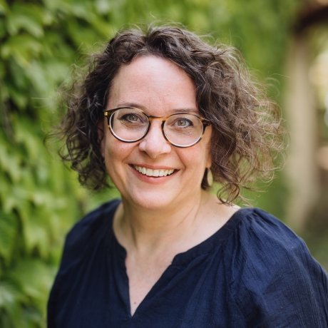 Woman with curly hair and glasses smiling, standing outdoors with greenery in the background.