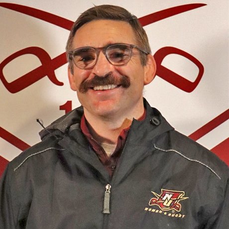 Man with mustache and glasses smiling in front of backdrop with crossed sabres.