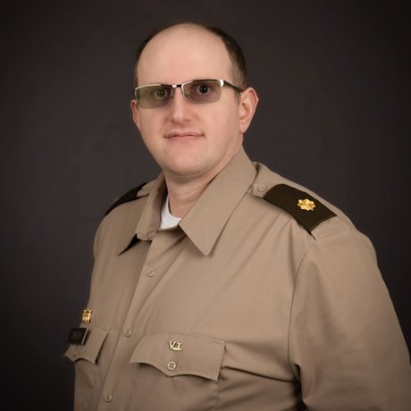 Person wearing a tan military uniform with dark shoulder epaulets and a gold oak leaf insignia, standing against a dark background.