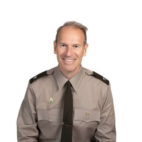 Individual wearing a tan military uniform shirt with dark brown shoulder epaulets, a dark brown tie, and two pins on the chest pockets against a white background.