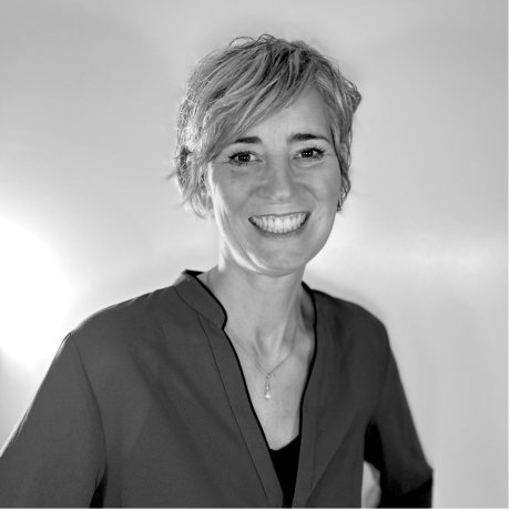 Individual with short, light hair wears a dark V-neck blouse and pendant necklace, standing in front of a plain light background in a black-and-white portrait.