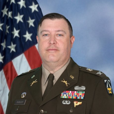 U.S. Army officer in dress uniform with name tag reading Dudley, various ribbons, badges, and insignia, standing in front of an American flag and blue backdrop.