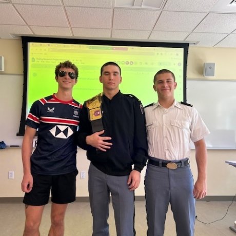 Three young men are standing together in a classroom, posing for a photo. The person on the left is wearing sunglasses and a sports jersey, the person in the middle is in a dark jacket with a patch on the shoulder, and the person on the right is wearing a white uniform shirt with gray trousers, typical of a military or cadet setting. A projection screen and whiteboards are visible in the background, suggesting this was taken during or after a class or presentation.