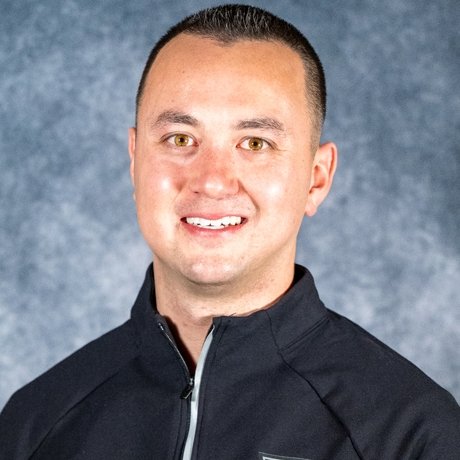 A person with short dark hair wears a dark zip-up jacket and smiles at the camera against a mottled gray background.