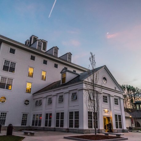 Large white building with square windows and lit rooms at dusk, with clear sky and trees in the background.