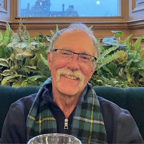 A man smiles while sitting indoors with plants behind him.