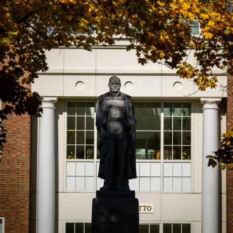 A statue of Alden Partridge stands in the foreground framed by autumn-colored leaves, with a classic building featuring white columns in the background.