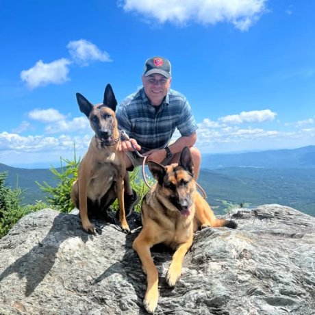 A man standing on a mountainside smiles, posing behind his two dogs.