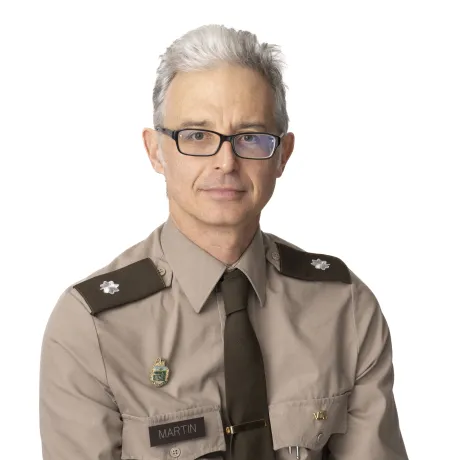 Person in a tan military-style uniform with shoulder insignia, name tag reading “MARTIN,” tie, and pins on the chest.