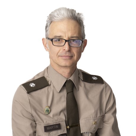 Person in a tan military-style uniform with shoulder insignia, name tag reading “MARTIN,” tie, and pins on the chest.