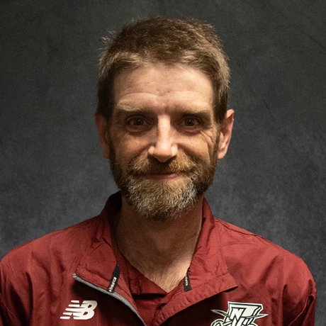 Head-and-shoulders portrait of a bearded man wearing a maroon athletic jacket, photographed against a dark neutral background.