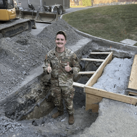 A person in military uniform stands in a shallow construction trench giving two thumbs up, with gravel piles, wooden forms, and excavation equipment nearby.