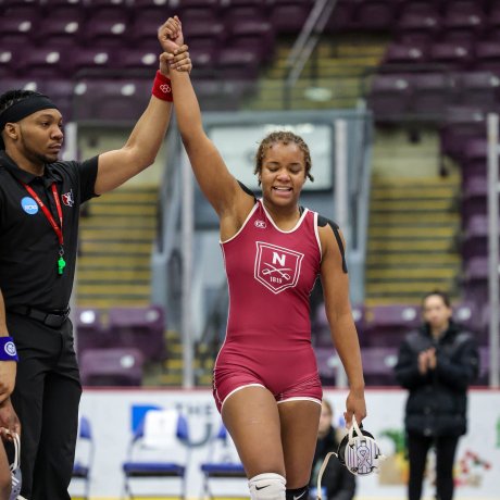 A wrestler in a maroon uniform stands on a mat as a referee lifts one arm in victory, with empty seats and a few people in the background.