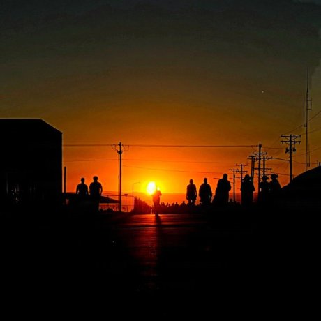 Silhouetted people walk along a road at sunrise with utility poles on both sides.
