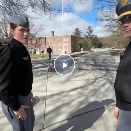 Two cadets stand on a campus walkway with a brick building in the background.