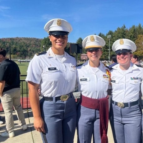 Four cadets in white dress uniforms stand together beside a football field on a sunny day.