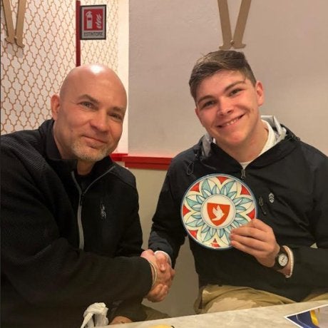 Two people sit at a table shaking hands, while one holds a circular artistic tile.