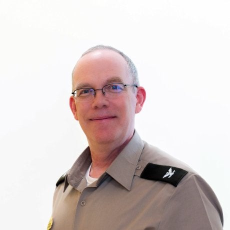 Person wearing glasses and a gray uniform shirt with shoulder insignia poses against a plain light background.