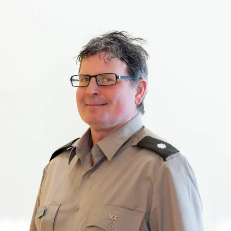 Person wearing glasses and a gray uniform shirt with shoulder insignia poses against a plain light background.