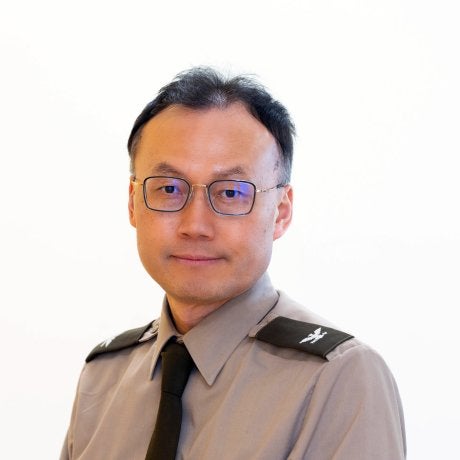 Person wearing glasses, a gray uniform shirt, and a black tie poses against a plain light background.