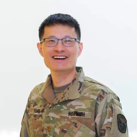 Person wearing glasses and a Norwich camouflage uniform faces the camera against a light background.