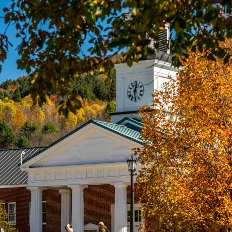 People walk past a brick building with white columns and a clock tower framed by autumn trees.
