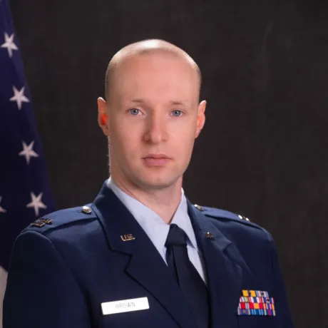 Uniformed service member stands in front of a U.S. flag and dark backdrop.