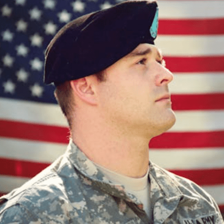 Service member in camouflage uniform and black beret faces right with an American flag in the background.