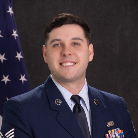 Service member in blue dress uniform stands in front of a U.S. flag and dark backdrop.