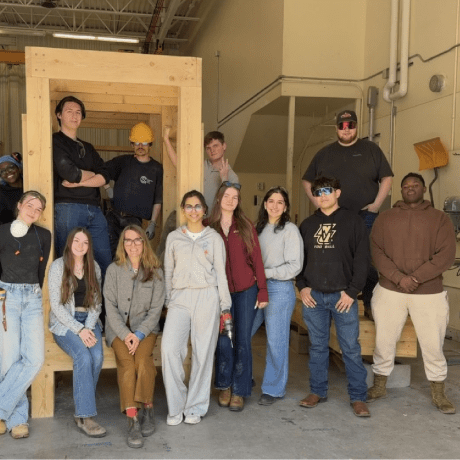 Students and an instructor pose beside a framed wood structure inside a construction lab