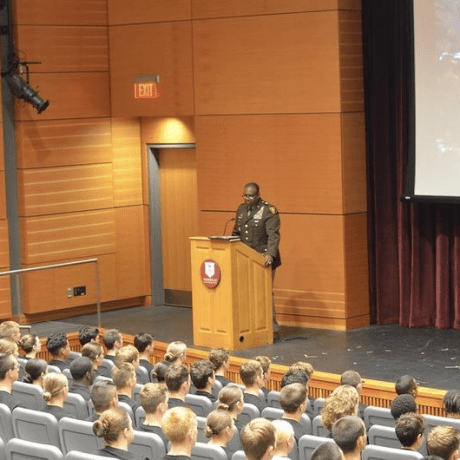 Service member speaks at a podium in an auditorium before an audience of seated cadets.