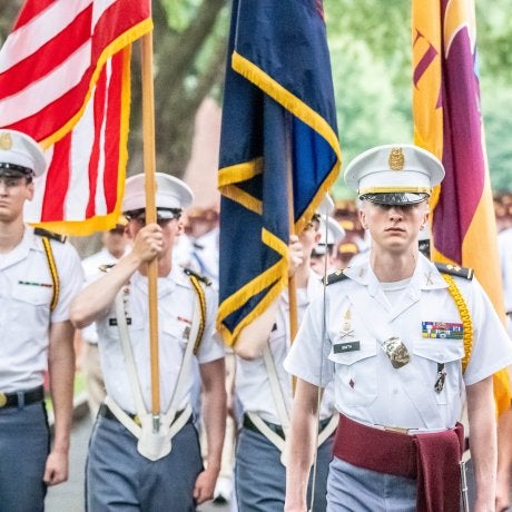 Cadets in uniform march in formation carrying the American flag and ceremonial flags during a parade.