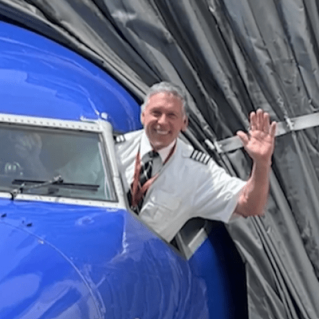 A pilot stands in the cockpit leaning out the open window, waving.