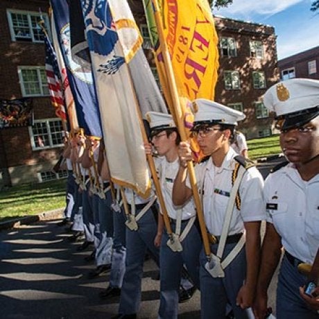 Norwich cadets stand in formation carrying flags and a rifle.
