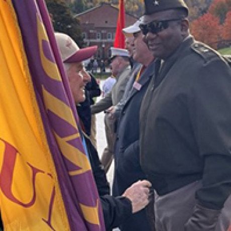 A person holding a yellow and purple flag speaks with a person in a military-style uniform at an outdoor gathering.