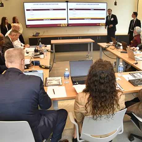Presenter speaks to attendees seated around tables as two screens display a legacy fleet presentation.