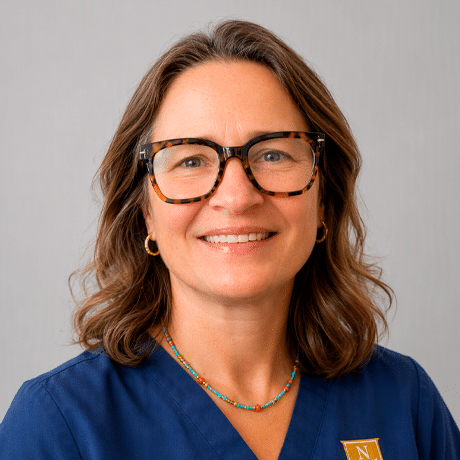 A person wearing glasses and blue Norwich University scrubs poses against a gray background.