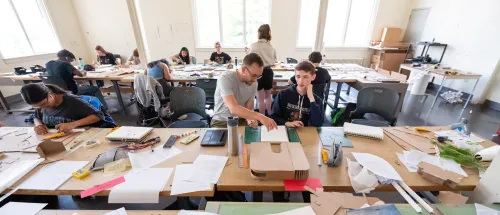 Students working on architectural models and drawings at large tables in a bright classroom at Norwich University.