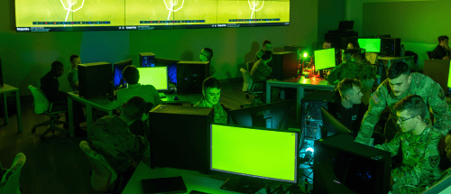 Students in military uniforms working at desktop computers in a green-lit Norwich University cybersecurity lab, with digital displays showing network diagrams.