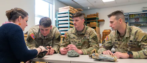 Three Norwich University cadets in camouflage uniforms examine rock and fossil specimens with an instructor in a campus geology lab.